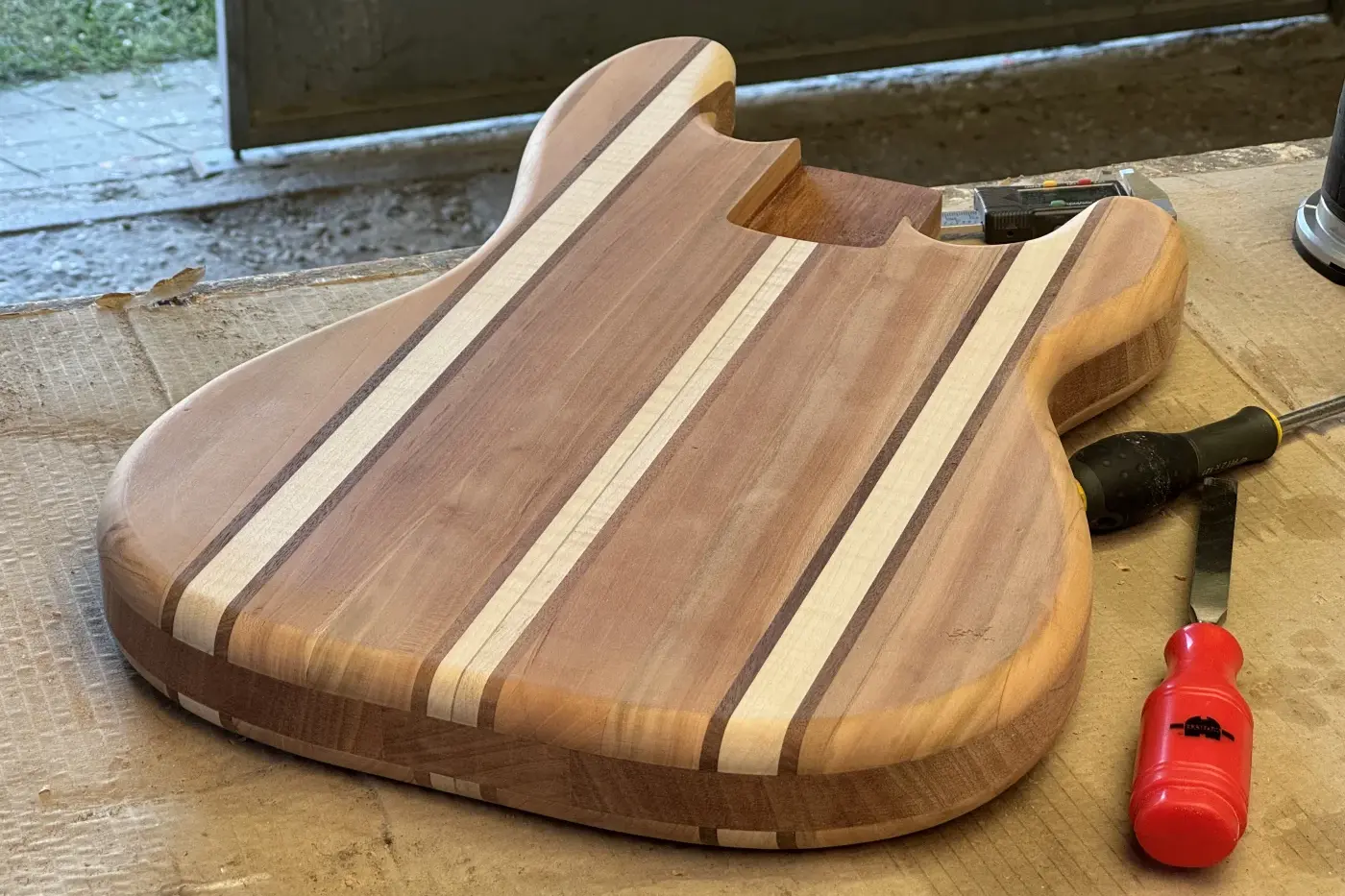 Vista dall'alto del body del Precision Bass laminato artigianale posato sul banco di lavoro con utensili, fronte con texture a strisce di legni misti