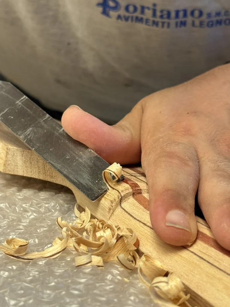 Primo piano dello scalpello in lavoro sulla voluta del manico multi-lamina del Precision Bass, con trucioli ricci di ramino sul piano