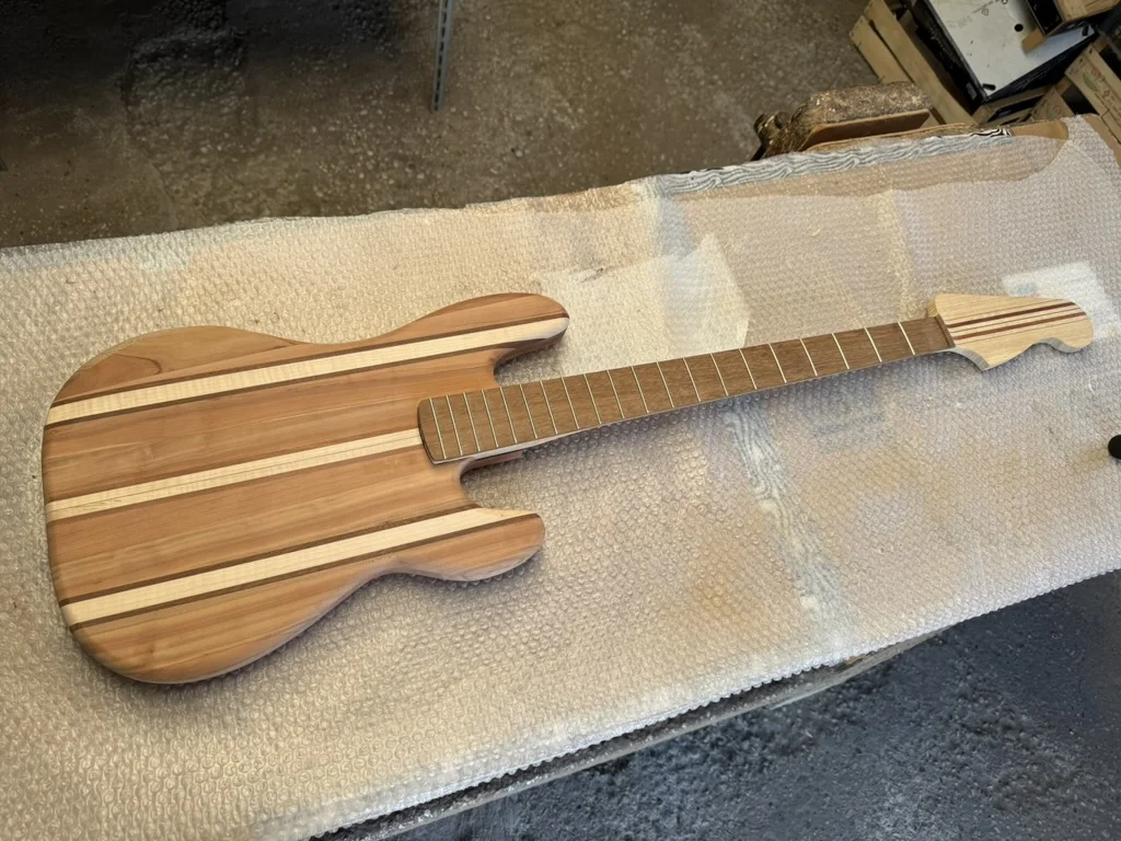 Vista frontale del Precision Bass artigianale multi-lamina: body a strisce di ramino e acero, manico sagomato con tastiera in iroko e tasti battuti, su piano di bolla